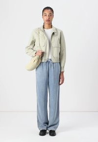 Veste vert clair avec des poignets élastiques et deux poches avant, t-shirt blanc, jean bleu à jambes larges et chaussures noires ; tenant un sac beige.