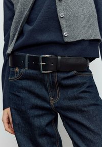 Cintura in pelle nera con fibbia in metallo argentato, indossata su jeans denim blu scuro con dettagli di cucitura arancione.