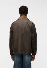 Άνδρας που φοράει ένα σκούρο καφέ κεριδωμένο μπουφάν με λινό γιακά από κορντουρέ και μπλε τζιν τζιν, εμφανιζόμενος από πίσω σε λευκό φόντο.