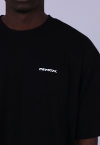 Černé bavlněné tričko s kulatým výstřihem, krátkými rukávy a bílým logem "CRYSTAL" na levé straně hrudi.