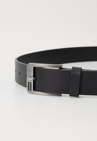 Ceinture en cuir noir avec une boucle en argent mat, présentant une forme rectangulaire, un petit logo et des trous ajustables pour un meilleur maintien.