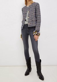 Giacca in tweed intrecciato multicolore blu e bianco, con bottoni dorati e rifiniture in pelliccia sulle maniche, abbinata a jeans grigio scuro e stivali neri.