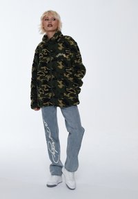 Giacca in fleece con motivo mimetico verde e marrone, chiusura frontale con zip, modello in piedi con jeans azzurri chiari e dettagli bianchi, oltre a sneakers bianche.
