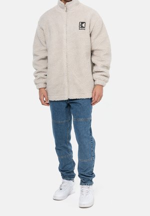 Chaqueta de forro polar de color beige claro con cremallera frontal, cuello alto y parche de logo negro. Combinada con jeans de mezclilla azul y zapatillas blancas.