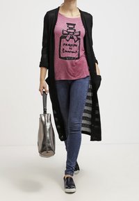 Černý dlouhý kardigan, růžové tričko s grafickým potiskem vůně, modré džíny, stříbrná taška, černé boty na klínku.