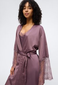 Γυναίκα με σγουρά μαλλιά που φοράει σατέν ρόμπα σε μωβ απόχρωση με μανίκια διακοσμημένα με δαντέλα, δεμένη στη μέση, πάνω από μια ταιριαστή καμισόλα.