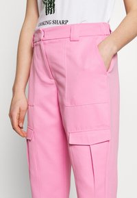 Różowe spodnie cargo o gładkiej teksturze, z przednimi kieszeniami, paskami na pasie i luźnym krojem. Stebnowane detale na kieszeniach i szwach.