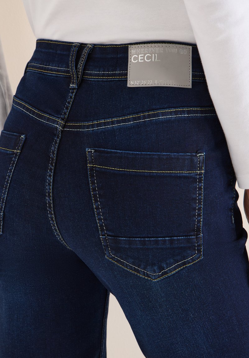 Jean en denim bleu foncé avec une coupe élégante, présentant des surpiqûres jaunes contrastantes et deux poches arrière. L'étiquette sur la ceinture indique "CECIL".