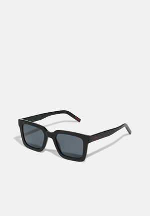 Occhiali da sole neri a forma rettangolare con lenti scure, montatura in plastica spessa e un accent di logo rosso sulla tempia. Design geometrico slanciato.