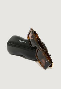 Sunglassi iz mačje dlake s kvadratnim okvirjem, rjavo obarvanimi lečami, zlatimi poudarki in črnim trdim etuijem s logotipom 'vogue'.