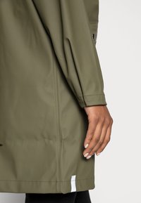 Alyvuogių žalios spalvos nepralaidus vandeniui švarkas su lygiu blizgančiu paviršiumi, ilgomis rankovėmis ir baltu etikete apačioje. Rankos delnas remiasi į audinį.