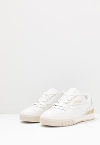Sneaker bianche in pelle con puntale perforato, design con lacci e accenti dorati a contrasto sul tallone. Presentano una suola piatta in gomma.