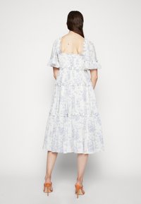 Kwiatowa sukienka midi z białym tłem i niebieskimi wzorami, z marszczoną górą, falbaniastymi rękawami i warstwową spódnicą. Widoczne pomarańczowe obcasy.