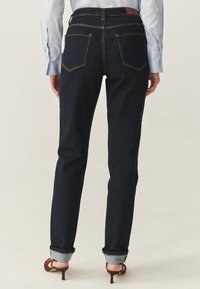 Jeans de mezclilla azul oscuro con un diseño de pierna recta, bolsillos traseros visibles, costuras amarillas y dobladillos remangados, combinados con tacones marrones.