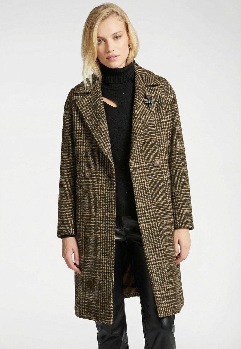 Manteau long marron à carreaux avec de larges revers, boutons croisés, et une silhouette cintrée, porté sur un col roulé noir et un pantalon en cuir.