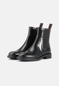 Botas Chelsea de cuero negro con paneles laterales elásticos, punta redondeada, tacón bajo y acento marrón en el elástico. Textura suave y diseño elegante.