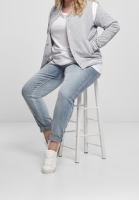 Woman wearing light blue jeans, white sneakers, white t-shirt, and gray jacket, seated on a white wooden stool against a plain background.