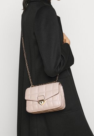 Sac en bandoulière rose matelassé avec un fermoir doré et une chaîne, présentant une forme rectangulaire et une texture lisse, porté sur un manteau noir.