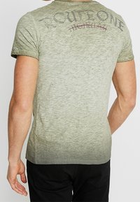 Grön gradient t-shirt med korta ärmar och rund hals. Har mörka bokstäver på ryggen och ett strukturerat, slitet utseende.