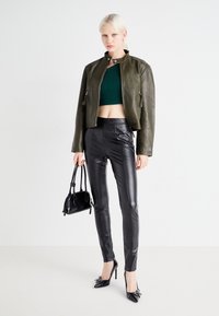 Top verde a maniche corte, giacca in pelle oliva, pantaloni in faux pelle neri, borsa nera e tacchi alti con un accento a fiocco.