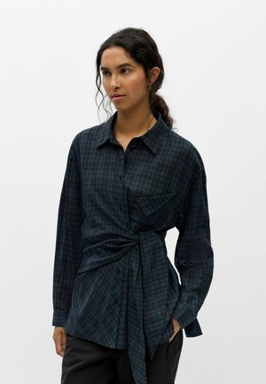 Jeune femme aux cheveux bouclés foncés portant une chemise à carreaux bleue et noire à manches longues nouée à la taille et un pantalon noir sur fond blanc.