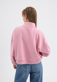 Roze sweatshirt met een hoge kraag, relaxte pasvorm en geribde zoom, gedragen met blauwe spijkerbroeken. De stof lijkt zacht en lichtjes gestructureerd.