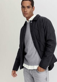 Schwarze gesteppte Jacke mit Knopfleiste, geformten Nähten und leichtem Glanz; über einem grauen Pullover und einem weißen Kragenhemd getragen.