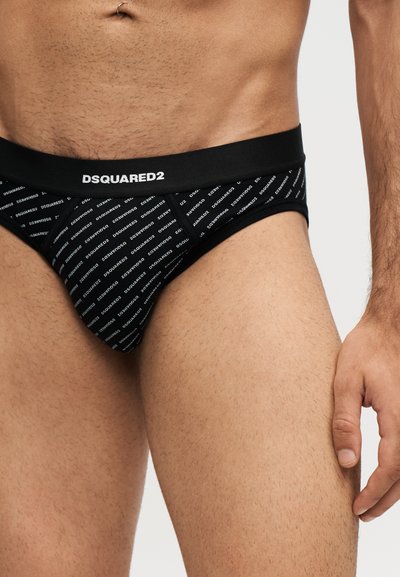 Torse masculin portant un slip noir DSQUARED2 avec un motif répétitif de logo blanc et un anneau de nombril visible.