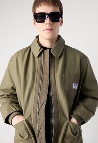 Khaki bomber jacket with a zip front, ribbed cuffs, chest pocket, and quilted lining. Black sunglasses with thick frames.