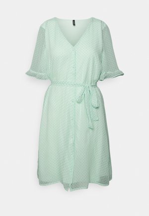 Robe vert clair jusqu'aux genoux avec un petit motif à pois, col en V, manches courtes à volants, boutons à l'avant et ceinture nouée à la taille.