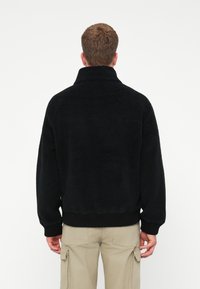 Man i svart fleecejacka och beige cargobyxor, stående med ryggen mot en enfärgad vit bakgrund.