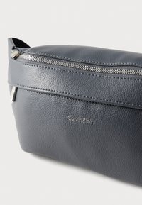 Γκρι δερμάτινη θήκη με ομαλή υφή, διαθέτει ασημένιο φερμουάρ και ένα μικρό ανάγλυφο λογότυπο. Συμπαγής, ορθογώνιο σχήμα.