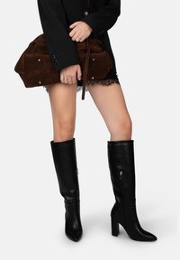 Botas de cuero negro hasta la rodilla con puntera puntiaguda y tacones en bloque. Un bolso de ante marrón con correa desmontable y cierres a presión.