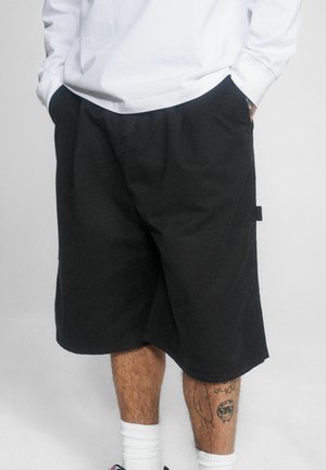 Osoba ubrana w czarne szorty sięgające kolan, białą koszulę z długim rękawem, białe skarpetki i widoczne tatuaże na nogach, z rękami w kieszeniach.