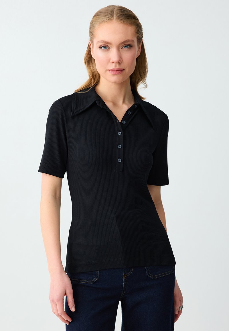 Femme aux cheveux blonds portant une chemise noire à manches courtes avec col et boutons, et un jean bleu foncé, sur un fond clair uni.