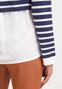 Navy and white striped sweater with a ribbed neckline, layered over a white blouse with a smooth texture and brown, fitted pants.