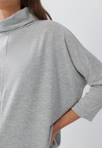 Grauer Sweatshirt mit Kapuze, lässiger Passform, Dreiviertelärmeln und sanfter Textur. Hat minimalen Nähten und eine Seitentasche.