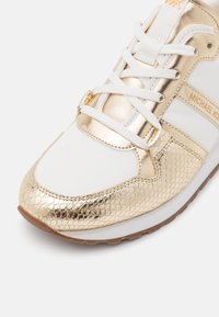 Zapatilla dorada y blanca con un acabado metálico texturizado, acentos de cuero suave, cordones blancos y suela de goma. Nombre de la marca visible.