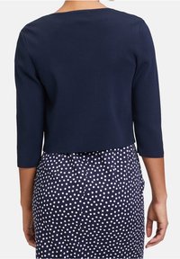 Pull court bleu marine à manches trois-quarts, en tissu tricoté texturé. Porté sur une jupe bleu marine avec un motif à pois blancs.