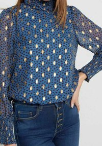 Blaue gemusterte Bluse mit goldenen und schwarzen Punkten, hohem Kragen, puffigen Ärmeln und gerafften Manschetten, kombiniert mit hoch taillierten Jeans.