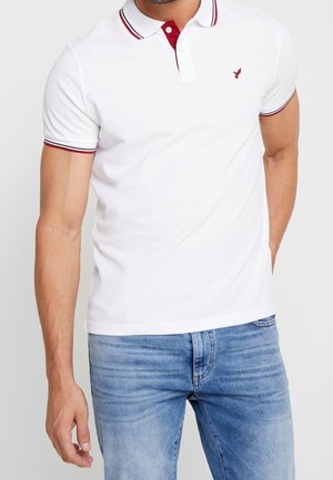 Polo shirt blanc à manches courtes, col rouge et bord-côte. Présente un petit logo rouge sur la poitrine. Matière en coton douce.