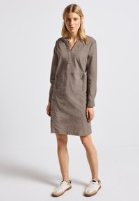 Khakifarbenes langärmliges Kleid mit V-Ausschnitt, zwei vorderen Reißverschlusstaschen und einer lässigen Passform. Getragen mit weißen Sneakern mit beigen Akzenten.
