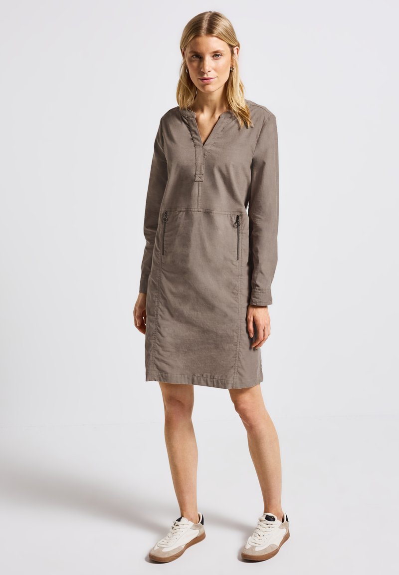 Khakifarbenes langärmliges Kleid mit V-Ausschnitt, zwei vorderen Reißverschlusstaschen und einer lässigen Passform. Getragen mit weißen Sneakern mit beigen Akzenten.