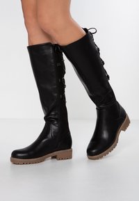 Nohy obuty v černých kožených holinkách s bočním zipem, šněrováním a silnými hnědými podrážkami na bílém podkladě.