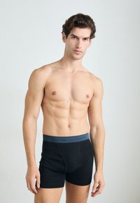 Schwarze Baumwoll-Boxershorts mit einem grauen Bund, der das Logo "Calvin Klein" trägt. Das Modell steht oberkörperfrei vor einem neutralen Hintergrund.