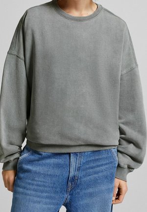 Osoba nosząca wyblakłą, szarą, oversizeową bluzę z prążkowanymi mankietami i dołem, w zestawieniu z jasnoniebieskimi dżinsami denim.