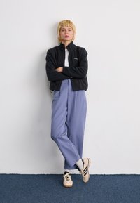 Black fleece jacket with a high collar, white t-shirt, lavender wide-leg trousers, and beige sneakers with black stripes, standing against a wall.