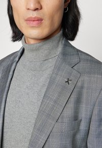 Blazer grigio a quadri con dolcevita abbinato. Presenta una spilla decorativa sulla rever e una trama liscia. Primo piano sulla parte superiore del corpo.
