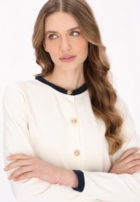 Cardigan crem cu guler bleumarin, trei butoni aurii și o textură moale, cu mâneci lungi și un design clasic.
