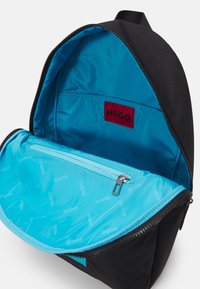 Sac à dos avec un extérieur noir et un intérieur bleu vif. Comprend une poche zippée et une étiquette logo rouge. Tissu lisse avec un design élégant.